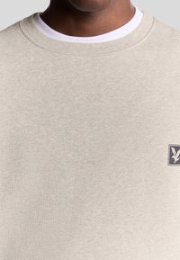 Felpa grigia chiara con collo a giro, texture di tessuto morbido e piccolo patch logo scuro con un design di aquila sul petto.