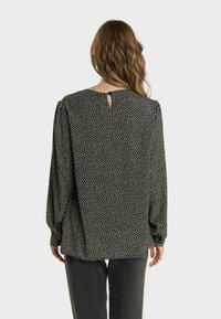 Blouse noire avec un motif à pois blancs, manches longues et un détail en forme de trou de serrure au dos. Fabriquée en tissu léger.