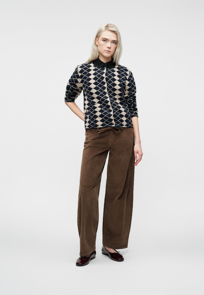Femme blonde se tenant avec une main derrière le dos, portant un pull à motifs losanges bleu marine et beige, un pantalon large marron et des chaussures sombres.