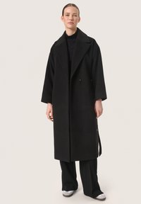 Cappotto di lana nero con ampio rever, doppi bottoni e maniche lunghe. Tessuto liscio e design lungo fino alla caviglia. Abbinato a pantaloni neri e scarpe grigie.