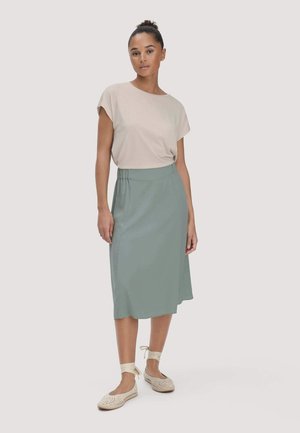 Vrouw met haar in een knot, gekleed in een beige top met korte mouwen, een groene midirok en beige veterschoenen, staand tegen een effen achtergrond.