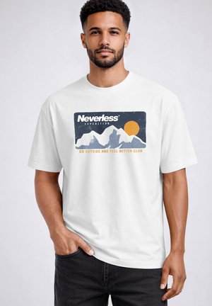 Mann mit kurzen Locken und Bart trägt weißes T-Shirt mit Berggrafik, orangefarbener Sonne und dem Text „Nevertheless Expedition, Go Outside and Feel Better Club.“
