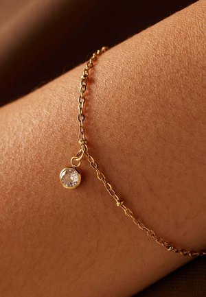 Gouden kettingarmband met een ronde heldere kristallen hanger, gepresenteerd tegen een huidkleurige achtergrond. De ketting is delicaat en fijn gekoppeld.