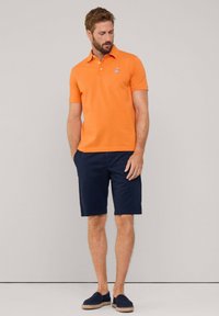 Polo arancione con colletto, maniche corte e un piccolo logo. Abbinato a pantaloni corti blu navy e scarpe slip-on blu navy con suola testurizzata.