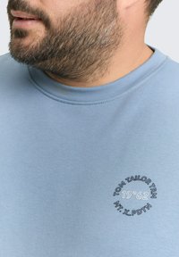 Close-up van de onderkant van het gezicht en de nek van een man die een lichtblauwe ronde hals T-shirt draagt met een zwart-wit cirkelvormig tekstlogo op de borst.