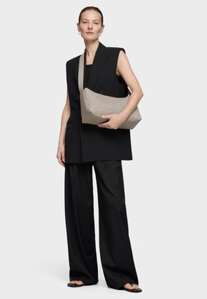 Kobieta ubrana w czarną marynarkę bez rękawów i szerokie spodnie, trzymająca taupe teksturowaną torebkę na ramię, stojąca na tle jasnego, gładkiego tła.