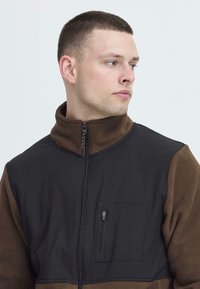 Svart og brun fleecejakke med zip og høy krage, glidelåslomme på brystet og strukturert overflate.