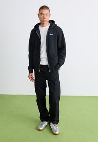 Mand står på grønne gulv iført sort zip-up hoodie, hvid t-shirt, sorte cargo bukser og sølvfarvede sneakers.