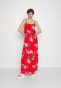 Vestido maxi vermelho com estampa floral, apresentando alças finas e uma cintura elástica. O material parece leve e tem um design fluido. Sandálias plataforma brancas.