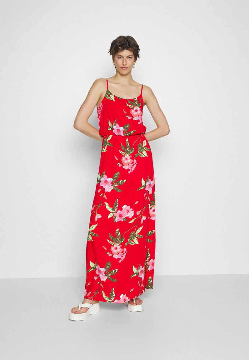 Vestido maxi vermelho com estampa floral, apresentando alças finas e uma cintura elástica. O material parece leve e tem um design fluido. Sandálias plataforma brancas.