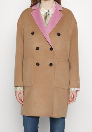 Kobieta ubrana w dwurzędowy płaszcz koloru wielbłądziego z różowym kołnierzem, na paskowanej koszuli zapinanej na guziki i niebieskich dżinsach.