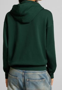 Hoodie verde scuro con cappuccio, polsini e orlo a coste. Tessuto morbido; presenta una vestibilità ampia. Indossato con jeans azzurri chiari.