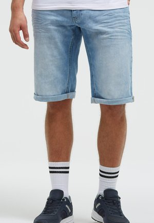 Mann, der hellblaue hochgekrempelte Jeansshorts, weiße Socken mit zwei schwarzen Streifen und dunkelblaue Turnschuhe auf weißem Hintergrund trägt.