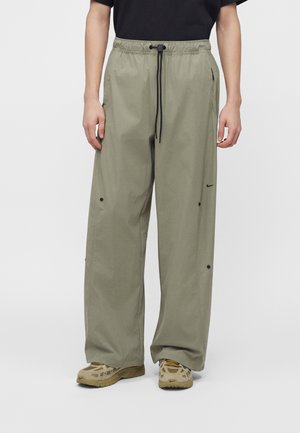 Personne portant un pantalon ample vert olive avec un cordon noir, des baskets beiges et un t-shirt noir à manches courtes, debout sur un fond uni.
