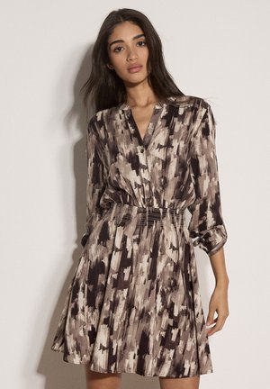 Femme aux longs cheveux foncés portant une robe à manches longues marron et beige à motif abstrait, avec des boutons et une taille élastique, debout contre un mur uni.