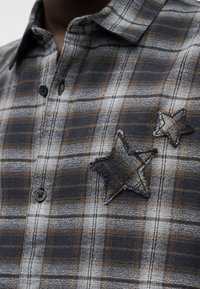 Γκρι και καφέ καρό πουκάμισο με κουμπιά, κατασκευασμένο από υφή βαμβακερό ύφασμα. Διαθέτει δύο κεντητά αστέρια στο αριστερό στήθος.