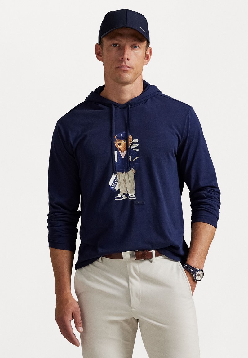 Man in een marineblauwe hoodie met teddybeer-afbeelding, lichtbeige broek, marineblauwe pet en marineblauwe horloge met 'Polo' band.