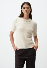 Pull crème à manches courtes en texture douce et duveteuse avec un col rond accentué de petits clous décoratifs en argent. Assorti à un pantalon sombre.