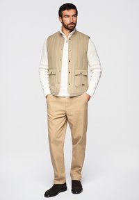 Hombre con chaleco acolchado beige sobre suéter blanco de punto trenzado, pantalones beige y zapatos marrón oscuro, de pie con las manos en los bolsillos.