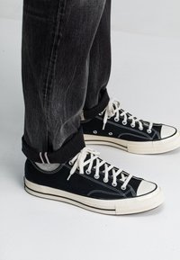 Schwarze Leinensneaker mit weißen Gummisohlen und Zehenkappe, ausgestattet mit weißen Schnürsenkeln und Ösen. Getragen mit grauen Jeans, umgeschlagen, um die Details zu zeigen.