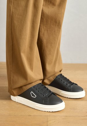 Pantofi sport negri din piele cu talpă din cauciuc albă și accente albe. Au un logo pe lateral și șireturi texturate. Poartați cu pantaloni bej.
