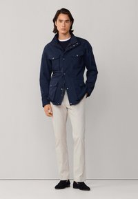 Chaqueta azul marino texturizada con cuello, dos bolsillos en el pecho y dos bolsillos inferiores, combinada con pantalones de color claro y zapatos oscuros.