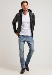 Giacca di pelle nera con cappuccio, t-shirt bianca, jeans azzurri chiari e stivali neri alla caviglia. Il modello sta in piedi con le mani in tasca.