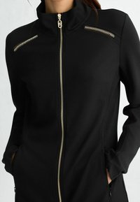 Czarna kurtka zapinana na zamek z wysokim kołnierzem, z złotymi elementami, kieszeniami na piersi na zamek oraz teksturowaną tkaniną po bokach.