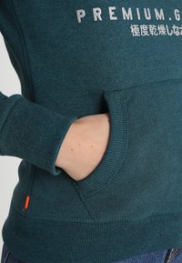 Felpa color turchese con tasca frontale, polsini e orlo a coste. Presenta del testo argentato sul petto e un piccolo etichetta arancione vicino all'orlo.