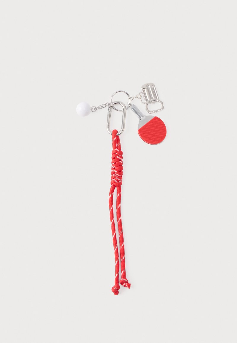 Porte-clés rouge avec cordon tressé, métal argenté, étiquette ronde rouge, clip et perle blanche. Présente une finition texturée sur l'étiquette.