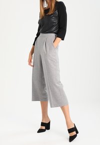Graue Wolle Culottes mit hoher Taille, Seitentaschen und Falten. Kombiniert mit einem schwarzen Kunstlederoberteil und schwarzen hohen Schuhen mit Riemen.