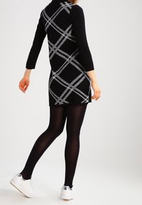 Czarna dzianinowa sukienka z białym geometrycznym wzorem, z długimi rękawami i wysokim dekoltem. Noszona z czarnymi rajstopami i białymi tenisówkami z metalicznymi akcentami.