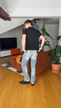 Nuori mies selinpäin, säätää vaaleansinisiä farkkujaan, seisoo puulattialla ruskean sohvan ja vihreän huonekasvin vieressä olohuoneessa.