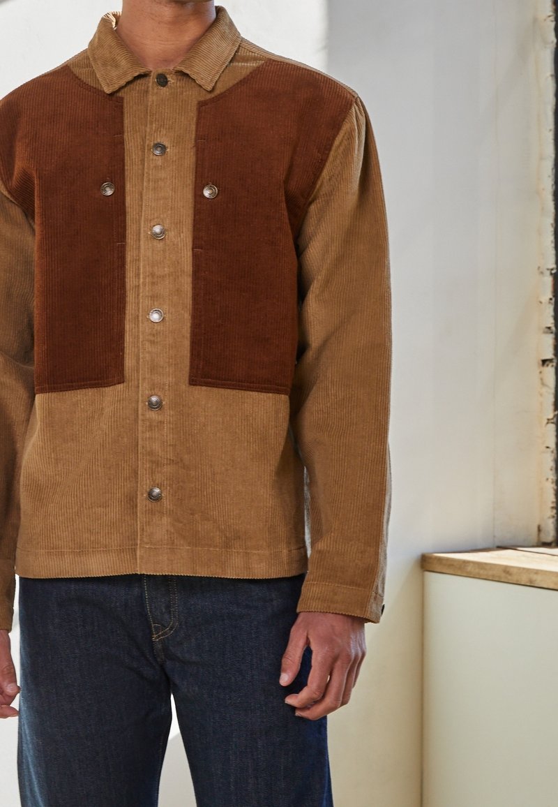 Homme portant une veste en velours côtelé boutonnée avec des panneaux marron foncé sur la poitrine et des manches beige, associée à un jean bleu foncé, debout à l'intérieur.