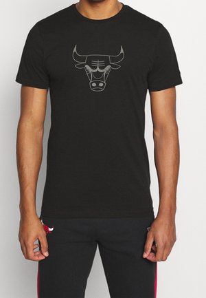 Mann trägt schwarzes T-Shirt mit stilisiertem Stierkopf-Logo und schwarze Hose mit kleinem rotem Stier-Logo am linken Oberschenkel.