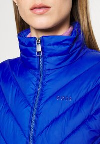 Blue padded jacket featuring a high collar, diagonal stitching, and a zip closure. The brand logo "BOSS" is visible on the left side.