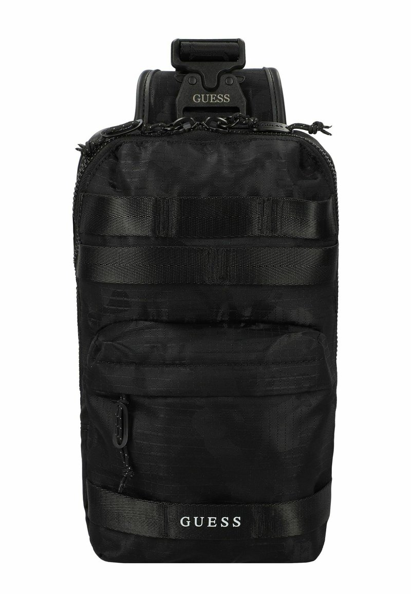 Zaino in nylon nero con superficie testurizzata, tasca frontale con zip e spallacci regolabili. Presenta il logo "GUESS" e webbing in stile tattico.