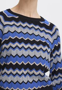 Pull tricoté présentant un motif en zigzag bleu, gris, noir et beige. Encolure ronde, bords côtelés et tissu texturé.