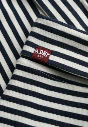 Gestreepte stof in marineblauw en wit, met een rood geborduurd etiket met "S. DRY NYC" op de kraag. Zachte brei-textuur.