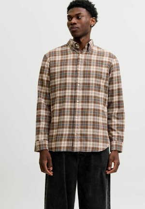 Homme portant une chemise à carreaux marron et beige boutonnée et un pantalon noir ample, debout avec les mains détendues le long du corps.