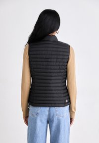 Črna, prešita jakna brez rokavov z visokim ovratnikom, oblečena čez svetlo rjav dolg rokav top, v kombinaciji s svetlo modrimi, ohlapnimi kavbojkami.