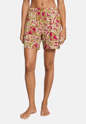 Jambes de femme portant un short beige avec un motif abstrait rose et marron, debout pieds nus devant un fond uni.