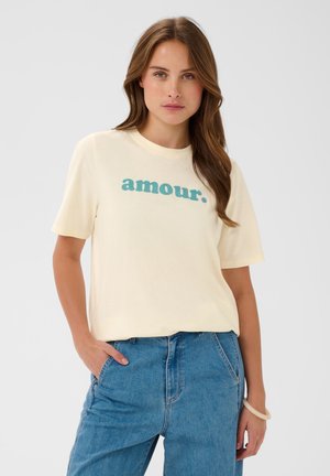 Kobieta z długimi brązowymi włosami ma na sobie kremowy t-shirt z napisem „amour.” na niebiesko i niebieskie dżinsy, stoi z jedną ręką w kieszeni.