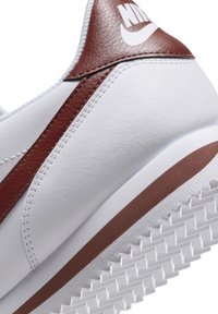 White leather sneaker featuring a brown accent stripe, textured brown heel, and a rubber sole with a serrated pattern. Nike logo visible.