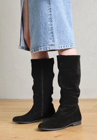 Persona que lleva botas de ante negras hasta la rodilla y una falda de mezclilla azul claro con una apertura en la parte delantera, de pie sobre un suelo de madera.