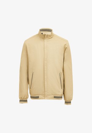 Beige takki, jossa on pystysuora kaulus, etuvetoketju, kaksi sivustavetoketjullista taskua ja raidallinen hihansuu. Valmistettu sileästä, kevyestä kankaasta.