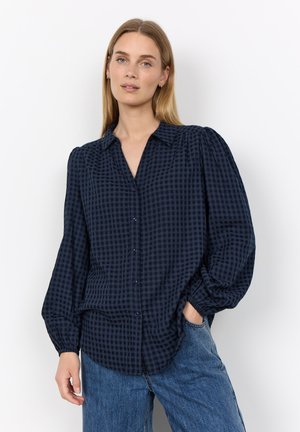 Woman with long blonde hair wearing a navy blue checkered button-up blouse and blue jeans, standing with one hand in pocket.