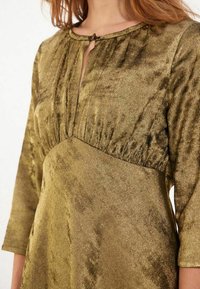 Robe vert olive texturée avec un décolleté en trou de serrure, des manches trois-quarts et un détail de taille froncée, fabriquée en matériau scintillant.