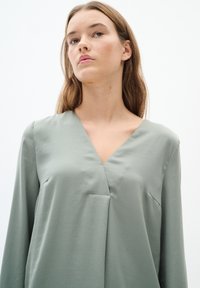 Maglietta verde chiaro a maniche lunghe con scollatura a V e una tasca frontale discreta. Tessuto morbido e vestibilità rilassata, dettagli di cucitura minimi.