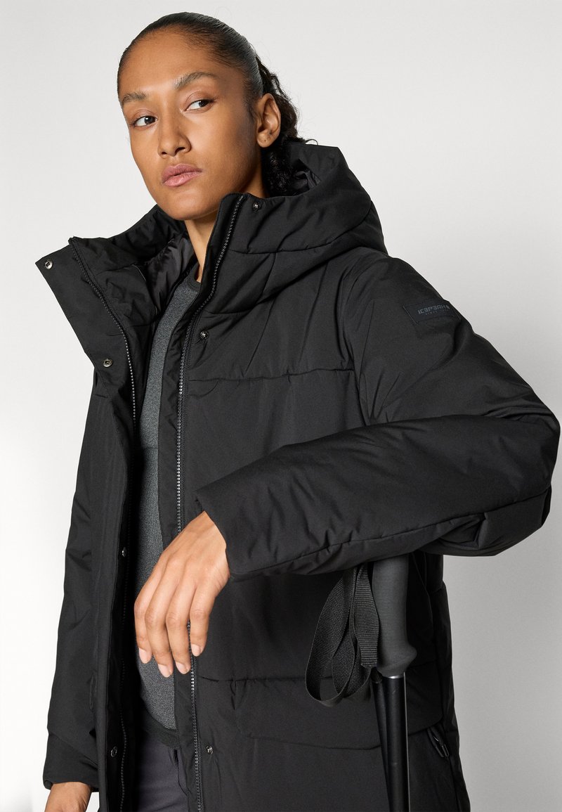 Frau in einer schwarzen Daunenjacke mit Kapuze, die einen schwarzen Trekkingstock vor einem schlichten hellen Hintergrund hält.